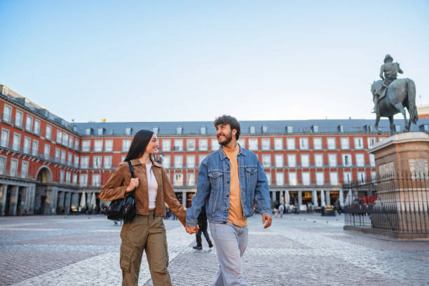 Hispanic couple explore Plaza Mayor in Madrid, walking hand in hand. The iconic square and its surroundings bring joy and togetherness as they enjoy their travel experience.