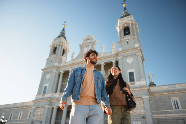 Hispanic couple holding hands and enjoying their visit to Almudena Cathedral in Madrid. They are smiling and showcasing joy and togetherness. Clear blue sky and historic architecture in the background.
