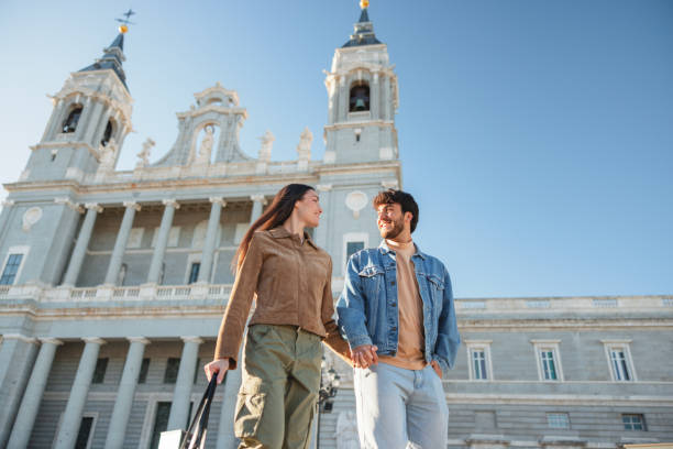 Hispanic couple visiting Madrid, holding hands and smiling in front of Almudena Cathedral. The image captures their joy and the beauty of the historic architecture.