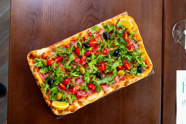 Rectangular pizza served on wooden board in fine dining, bacon, cherry tomato, black and green olives and arugula topping