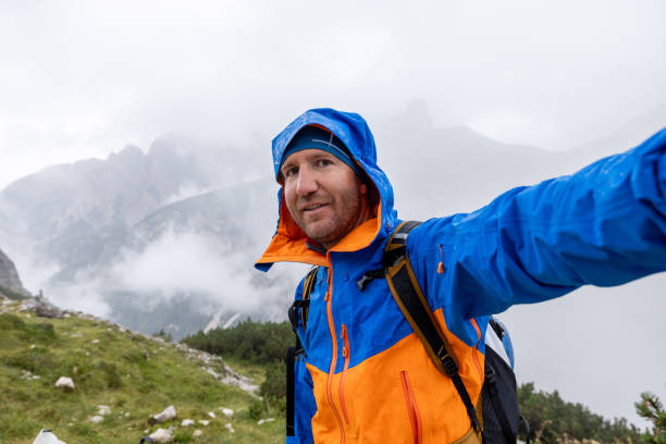 Hiker in blue and orange rain jacket taking a selfie on a misty mountain trail. The cloudy landscape creates a serene and adventurous atmosphere.