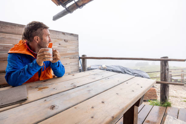 Male hiker sits at a rustic wooden table in a mountain cabin, savoring a hot drink while surrounded by foggy, serene mountain scenery. A peaceful moment after a hike.