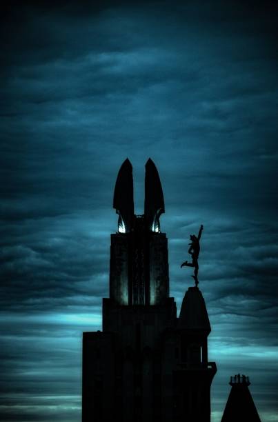 Silhouette of a tall building with a statue on top against a dramatic cloudy sky at dusk, showcasing architectural details and moody atmosphere.
