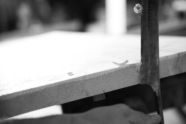 Carpenter is using a hand plane on a wooden plank in a workshop, with wood shavings visible on the surface. - black n white -