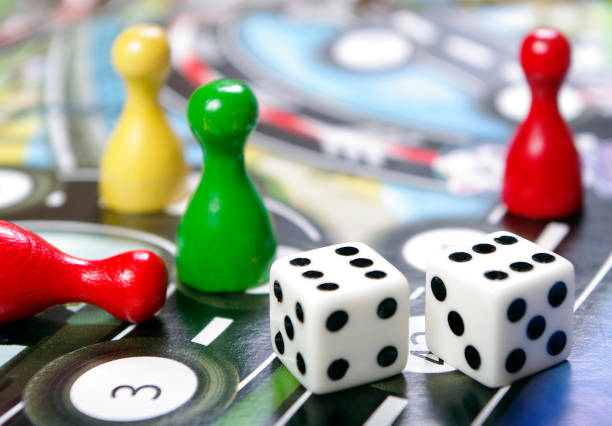 A high-angle view of four friends sitting around a glass table in a cozy living room in the North East of England, engaged in playing Snakes and Ladders They are having fun, smiling and laughing. One member of the group is taking his turn. The group are dressed casually, with warm lighting creating a relaxed atmosphere. The living room has a modern yet cozy feel, with a beige sofa, and a leopard-print cushion,  Various game pieces, cards, and tokens are scattered on the table as they enjoy their game night together.