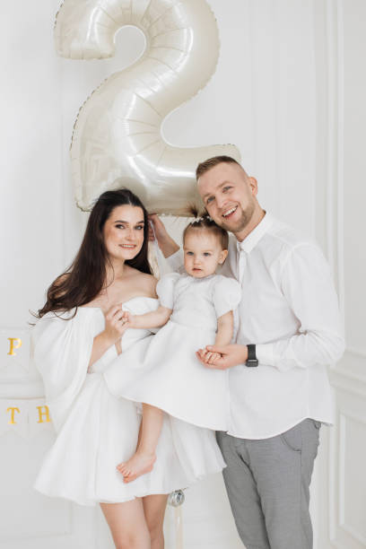 Caucasian family with young parents and daughter celebrating second birthday. Parents wearing white and grey outfits, daughter in white dress. Joyful atmosphere with large balloon shaped as number two