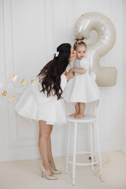 Bride mother kissing young daughter celebrating second birthday. White, feminine-themed attire adds elegance. Scene features mother, daughter with large balloon in stylish, minimalistic setting.
