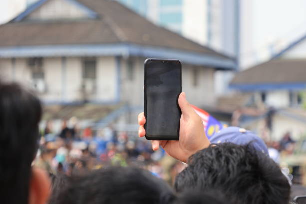 A hand holding a smartphone capturing the vibrant Malaysia Independence Day Parade, with blurred spectators in the background. Perfect for showcasing modern event photography and national celebrations.
