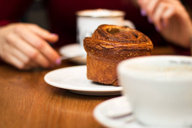 Cinnamon bun in focus with two coffees and a set of hands blurred in the background