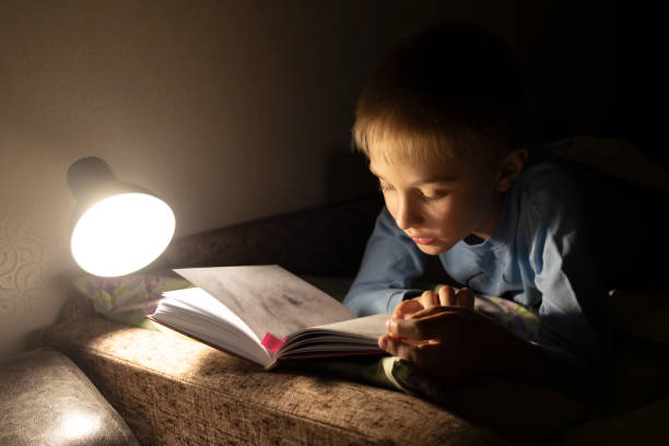 istock 9-year-old boy is reading book with interest while lying in bed in evening by light of lamp. Booklover 2176166597