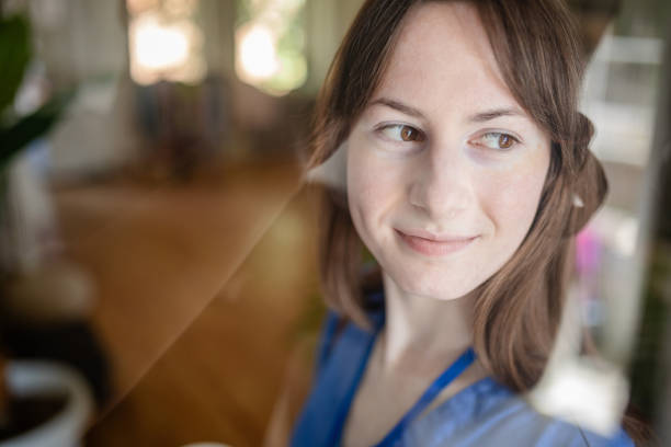 A nurse in blue scrubs stands by the window smiling with contentment, suggesting a moment of calm and reflection amidst the busy healthcare environment.