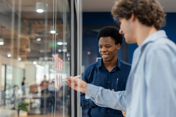 istock Multiracial business development team in a modern meeting room, sharing ideas and brainstorming by writing on sticky notes and attaching them to the glass. The session emphasizes teamwork, creativity, and strategic thinking for business improvement. 2176173673