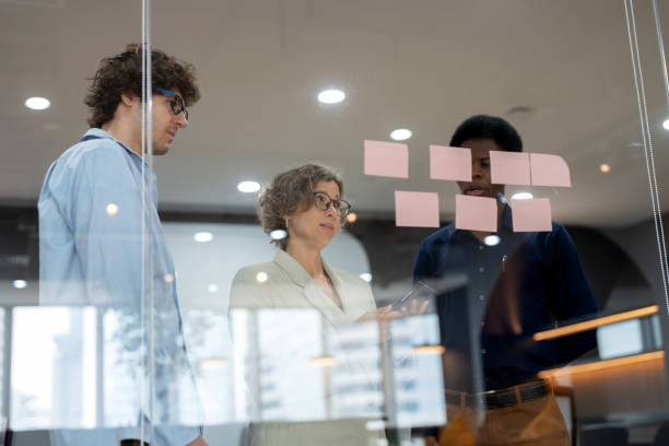istock Diverse group of business development brainstorming product development ideas in a meeting room, using sticky notes on a glass wall. Collaboration, creativity, and teamwork drive innovation, business strategy, and digital transformation in a modern office 2176173681