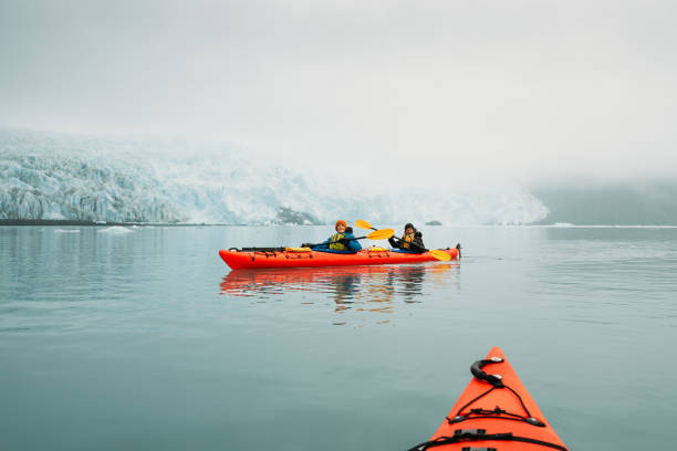 View of dock, boat and several kayaks next to wooded shore off Hoonah, Alaska, USA, with snowcapped mountain range covered with clouds in background