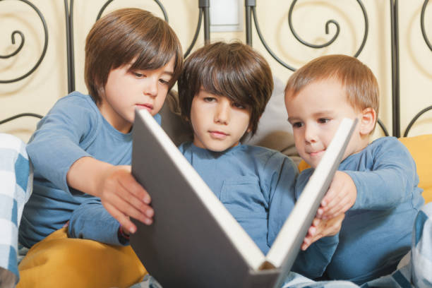 Three young boys are sitting together on a bed, focused on reading a large book. Dressed in matching blue pajamas, they appear deeply engaged in the story, sharing a quiet and intimate moment indoors.