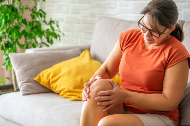 istock Woman suffering from knee pain. Massaging her knee with both hands sitting on sofa 2176187417