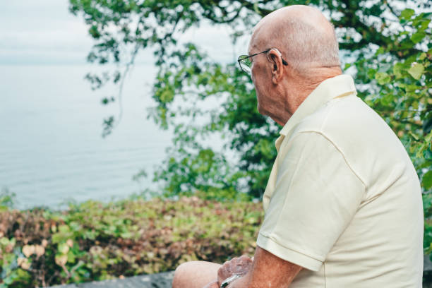 Portrait of an elderly man with bald head wearing a hearing aid sitting outdoors while looking towards the lake. Relaxed retired lifestyle