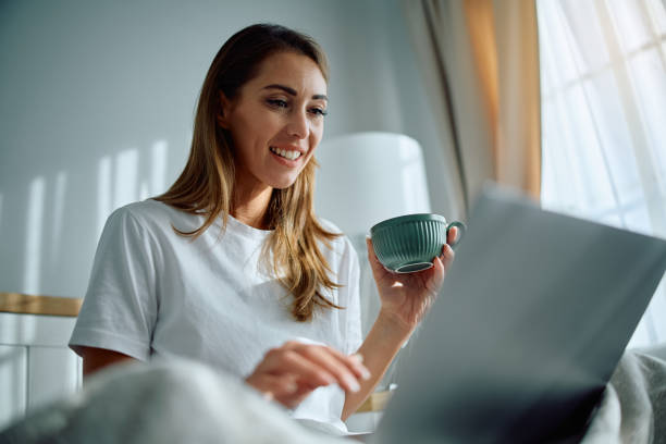 istock Happy woman drinking morning coffee while using laptop in bedroom. 2176214839