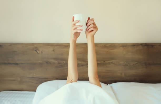 istock Cheerful lazy woman waking up after sleeping lying in bed holding cup in her hands asks for coffee 2176246095