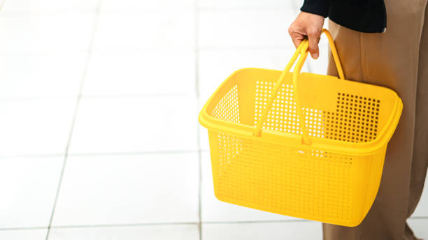 A person holding a bright yellow shewpping basket while walking on a tiled floor. The basket is empty and has a perforated design, indicating it's suitable for grocery shew