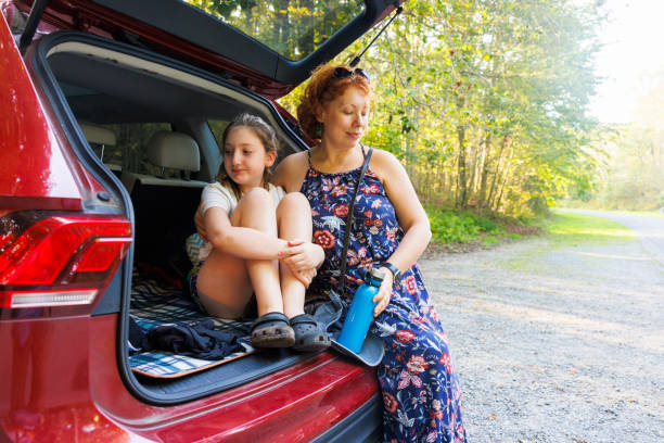 istock Pit stop during scenic drive. Mother and daughter relax on SUV trunk in forest setting. 2176297651
