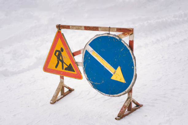 A construction warning sign and a detour sign are placed on a snow-covered road, alerting drivers to upcoming road work and guiding them to alternate routes.