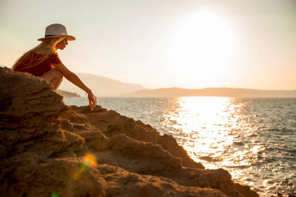 Beautiful teenage girl spending some time at the coastline cliff looking at sunset over the sea water. Young female tourist with hat relaxing by the ocean. Side view, copy space