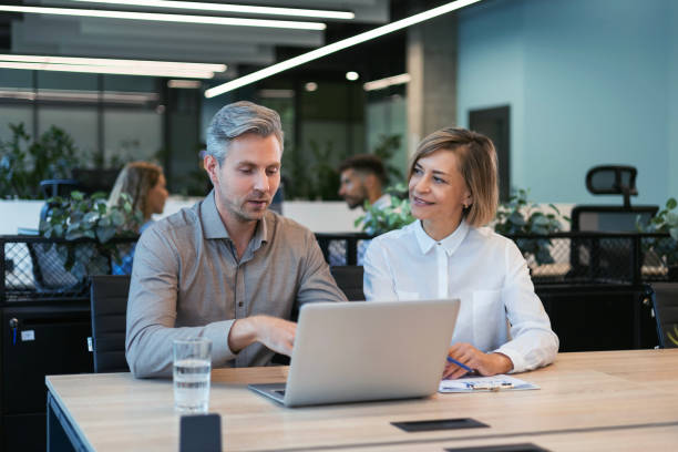 istock Two business people using laptop on desk discussing strategy company with other employees on background 2176392721