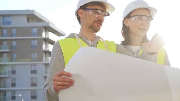 istock Two architects or engineers wearing safety hard hats and vests holding blueprint and discussing construction site project at sunrise, front view. Architecture, engineering and teamwork concept 2176393672