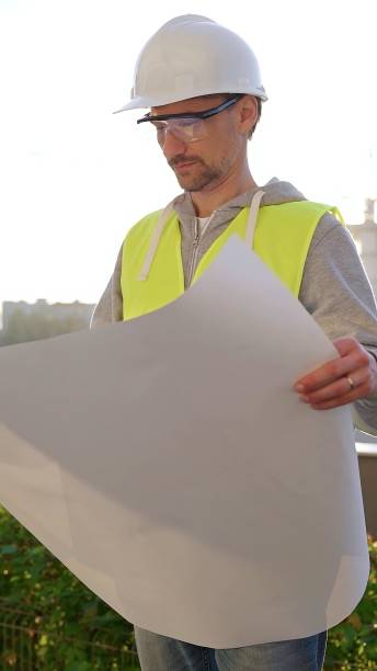 istock Man an architect or engineer wearing safety hard hat and vest holding blueprint and looking to construction site at sunrise, front vertical view. Architecture concept 2176397628
