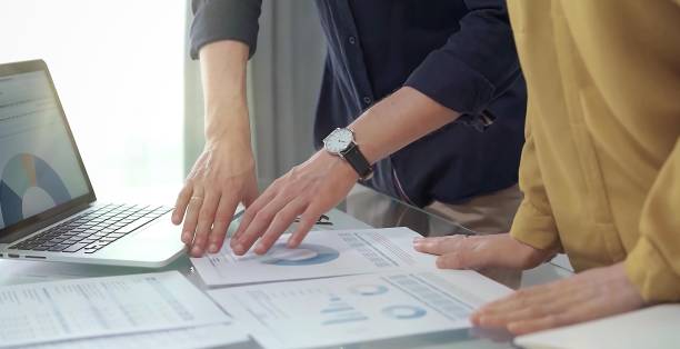 istock Business people, professionals analyzing financial charts and data on a desk while standing and using laptop computer in office. Dark blue shirt and yellow blouse 2176404914