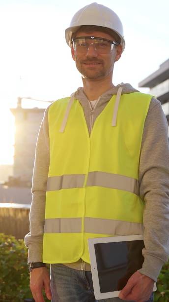 istock Construction site engineer holding tablet and wearing hard hat and safety vest with a sunrise at the background. Vertical portrait, looking at camera 2176408401
