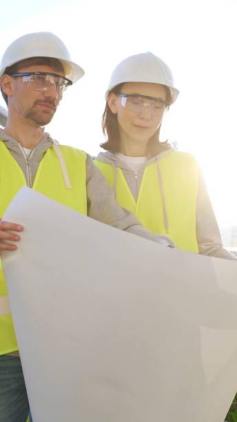 istock Two architects wearing safety hard hats and vests holding blueprint and discussing something on construction site at sunrise, front vertical view. Architecture and engineering concepts 2176431499