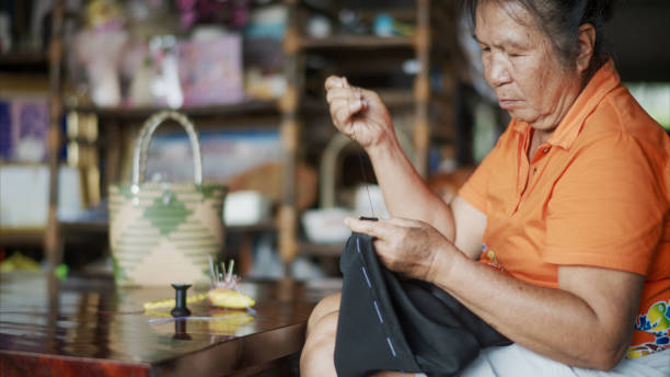 An elderly woman, is seen meticulously sewing fabric by hand. Her focus and precision emphasize the care and expertise involved in traditional craftsmanship. A woven bag and other crafting tools are visible, reinforcing the atmosphere of dedication to manual work. The essence of preserving cultural heritage through handcraft, with an intimate look at the art of sewing, passed down through generations.
