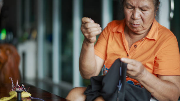 An elderly woman, is seen meticulously sewing fabric by hand. Her focus and precision emphasize the care and expertise involved in traditional craftsmanship. A woven bag and other crafting tools are visible, reinforcing the atmosphere of dedication to manual work. The essence of preserving cultural heritage through handcraft, with an intimate look at the art of sewing, passed down through generations.