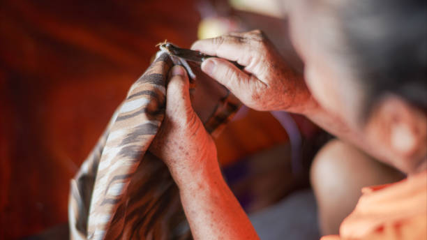 An elderly woman, is seen meticulously sewing fabric by hand. Her focus and precision emphasize the care and expertise involved in traditional craftsmanship. A woven bag and other crafting tools are visible, reinforcing the atmosphere of dedication to manual work. The essence of preserving cultural heritage through handcraft, with an intimate look at the art of sewing, passed down through generations.