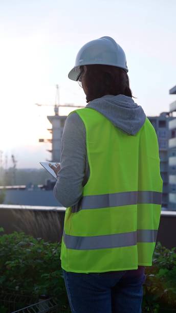 istock Female construction engineer with a white hard hat and safety vest is using digital tablet while inspecting a construction site in the early morning at sunrise, back view. Engineering concept 2176451182