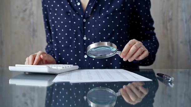 istock Auditor woman analyzing financial documents with magnifying glass and calculator at her office desk in formal dark blue blouse with polka dots. Business people concept 2176454053