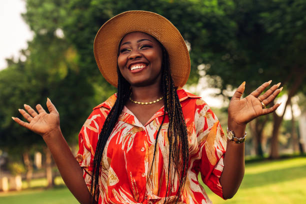 A young woman stands amidst a lush green backdrop, her wide smile and open hands radiating joy and warmth. She wears a straw hat and a bright, patterned dress, adding vibrancy to the scene. The soft sunlight filtering through the trees highlights her cheerful demeanor, capturing a carefree and festive spirit. The natural setting enhances the sense of freedom and positivity, while her expressive pose and genuine laughter invite the viewer to share in this moment of pure happiness. Her outfit, combined with her joyful expression, symbolizes a celebration of individuality, confidence, and the simple joys of life.