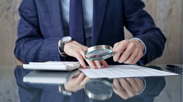 istock Auditor man analyzing financial documents with magnifying glass and calculator at his glass office desk in formal blue jacket. Business people concept 2176457601