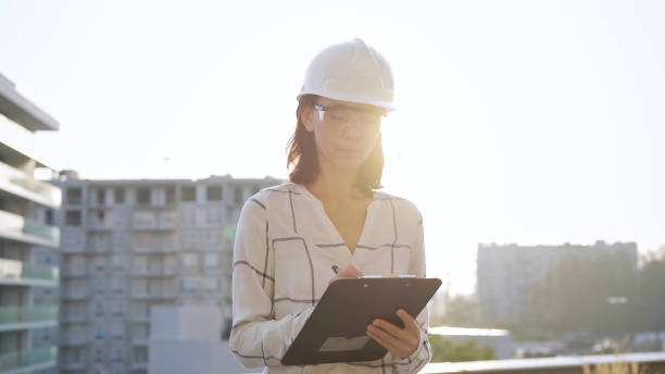 istock Woman construction engineer wearing white checked blouse and hard hat is making notes on a clipboard while inspecting a building site at sunset, front view. Architecture and engineering concepts 2176464299