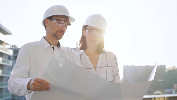 istock Two architects, wearing safety helmets and glasses, are examining blueprints at a construction site during sunrise or sunset, front view. Architecture and engineering concepts 2176464756