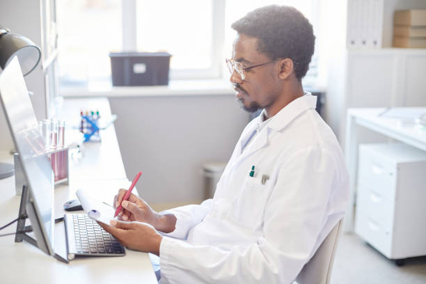 istock Male Chemist Writing in Notepad Sitting at Table Equipped with Computer in Laboratory 2176475723