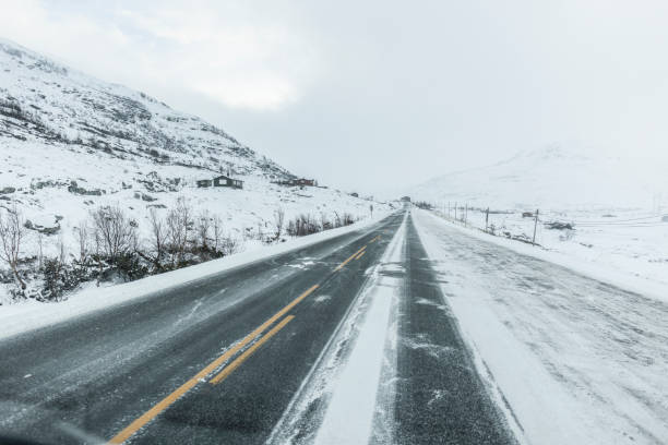 An empty snow-covered road stretches through a remote mountainous landscape during winter, surrounded by snow and rugged terrain.