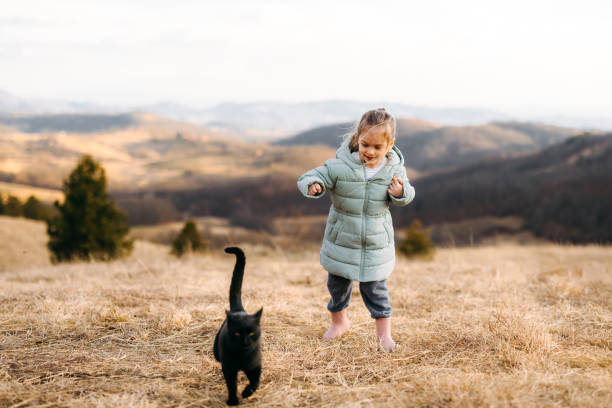 Chereful little girl play with two cats at countryside outdoor. People love pets. A beautiful girl gently hugs and caress her beloved cat while being outside in nature.