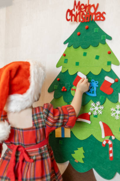 istock A young child decorates a colorful felt Christmas tree with ornaments while wearing a festive red hat in a cozy indoor setting 2176542455