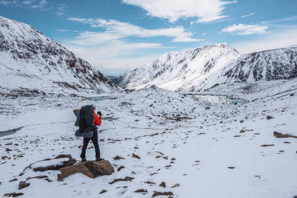 Man in red jacket with big backpack in snowy valley with alpine lakes under clouds in blue sky. Backpacker in snow-covered large mountains among glacial lakes, pure white snow and stones in sunny day.