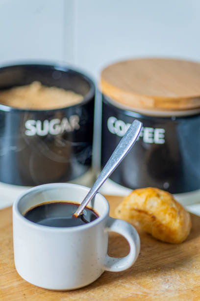 Freshly poured,hot,ready to drink,in an espresso coffee mug and silver spoon used to stir,on a wooden chopping board next to a bread roll,sugar and coffee containers out of focus in the background.