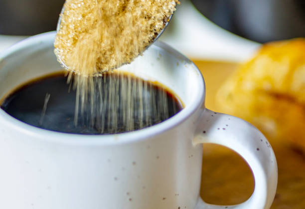 Demerara sugar falling from a silver teaspoon into a small cup of hot espresso coffee,in a domestic kitchen,Worcestershire,United Kingdom.