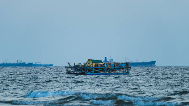 A Wooden Fishing Platform sits on the calm waters of the ocean, with larger cargo ships visible in the distance..
